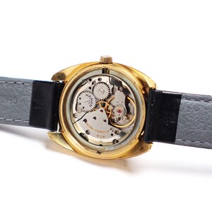 May include: Close-up of a vintage gold-toned wristwatch with a visible mechanical movement. The watch has a black leather strap and a gray leather backing. The watch face is visible, with the brand name "MARVIN" printed on it.