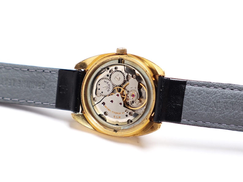 May include: Close-up of a vintage gold-toned wristwatch with a visible mechanical movement. The watch has a black leather strap and a gray leather backing. The watch face is visible, with the brand name "MARVIN" printed on it.