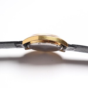 May include: A gold-toned wristwatch with a black leather band. The watch face is visible, and the band is attached to the watch case. The watch is set against a white background.