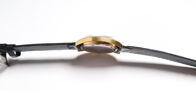 May include: A gold-toned wristwatch with a black leather band. The watch face is visible, and the band is attached to the watch case. The watch is set against a white background.