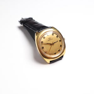 May include: A vintage gold-toned Marvin wristwatch with a black leather band. The watch face is gold with black hour markers and hands. The watch is angled slightly, showcasing its design and the brand name.