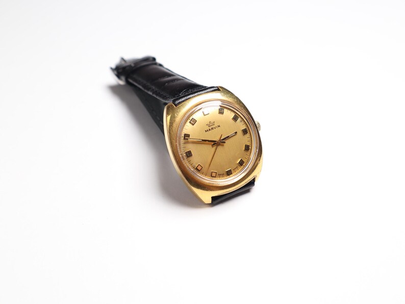 May include: A vintage gold-toned Marvin wristwatch with a black leather band. The watch face is gold with black hour markers and hands. The watch is angled slightly, showcasing its design and the brand name.