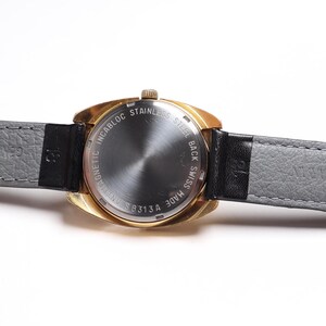 May include: A vintage gold-toned wristwatch with a gray leather strap and a black leather accent. The watch face is not visible. The back of the watch has text that reads "INCABLOC STAINLESS STEEL BACK SWISS MADE 313A".