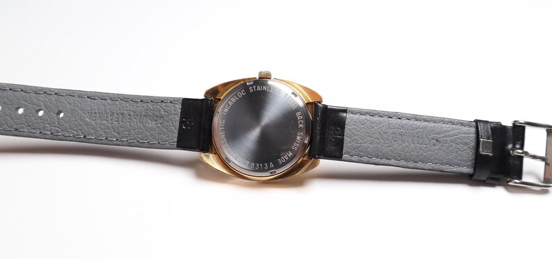 May include: A vintage gold-toned wristwatch with a gray leather strap and a black leather accent. The watch face is not visible. The back of the watch has text that reads "INCABLOC STAINLESS STEEL BACK SWISS MADE 313A".