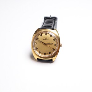 May include: A vintage gold-toned watch with a rectangular face and a black leather band. The watch face is gold with black markers and hands. The watch is labeled "MARVIN".