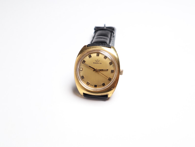 May include: A vintage gold-toned watch with a rectangular face and a black leather band. The watch face is gold with black markers and hands. The watch is labeled "MARVIN".