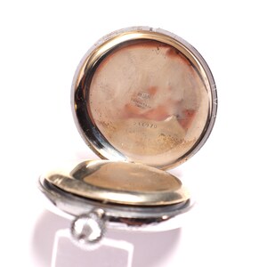 May include: Open pocket watch with a silver case and a gold-toned interior. The watch is partially open, revealing the inner workings and a small, textured surface. The watch has a small, silver winding stem.