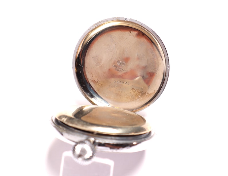 May include: Open pocket watch with a silver case and a gold-toned interior. The watch is partially open, revealing the inner workings and a small, textured surface. The watch has a small, silver winding stem.