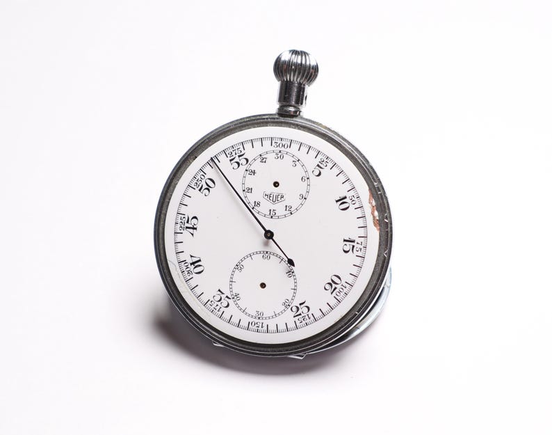 May include: A vintage Heuer stopwatch with a white face and black numerals. The stopwatch has a silver metal casing and a winding crown at the top. The face includes three sub-dials and the Heuer logo. The hands are black.