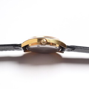 May include: A gold-toned wristwatch with a black leather band. The watch face is round, and the band is secured with a buckle. The watch is seen from the side, showcasing its profile.