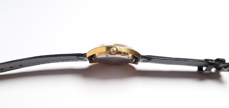 May include: A gold-toned wristwatch with a black leather band. The watch face is round, and the band is secured with a buckle. The watch is seen from the side, showcasing its profile.