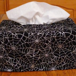 May include: A black tissue box cover with a silver spiderweb pattern. The cover is filled with white tissues.