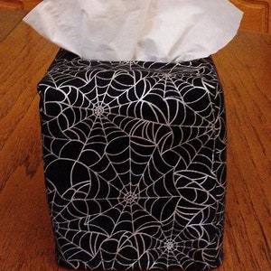 May include: A black tissue box cover with a white spiderweb design. The cover is made of fabric and has a square shape.
