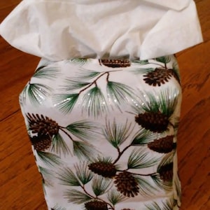 May include: A white tissue box cover with a pine cone and evergreen pattern. The cover is made of fabric and has a white tissue box inside.