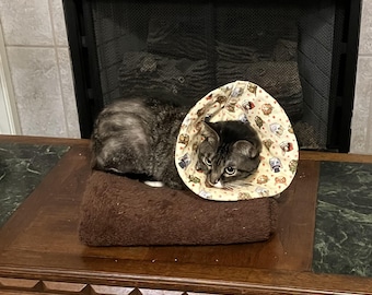 May include: A tabby cat wearing a beige and brown cone with a pattern of cartoon dogs and cats. The cat is sitting on a brown blanket on a wooden table.