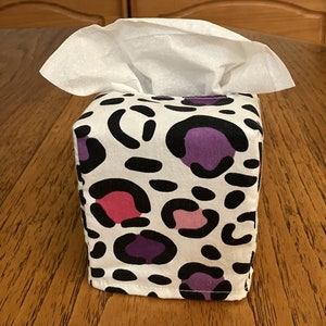 May include: A white tissue box cover with a leopard print design featuring black, pink, and purple spots.
