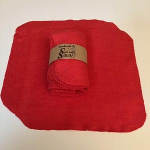 May include: A stack of red cloth napkins, with a rolled-up bundle secured by a brown paper band. The band reads "Handmade By Sew with Sarah!" in white script. The napkins are a solid, vibrant red color.