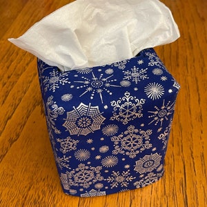May include: A blue tissue box cover with a silver snowflake pattern. White tissues are visible at the top. The box is square-shaped and sits on a wooden surface.