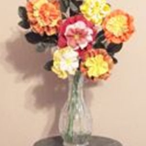 May include: A bouquet of colorful paper flowers in shades of orange, yellow, pink, and white arranged in a clear glass vase. The vase is sitting on a green and brown marble table.
