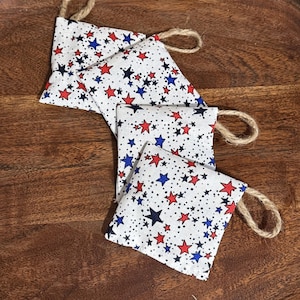 May include: Four small, square fabric sachets with a white background and a pattern of red and blue stars. Each sachet has a loop of natural twine attached. The sachets are arranged on a dark wooden surface.