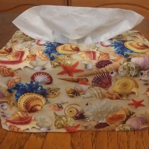 May include: A beige tissue box cover with a seashell design. The cover features a variety of seashells, starfish, and other ocean-themed imagery in shades of red, blue, yellow, and brown.