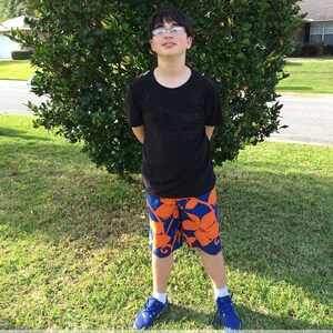 May include: A young person wearing a black t-shirt and blue and orange swim trunks with a floral pattern. The person is standing in front of a green bush.
