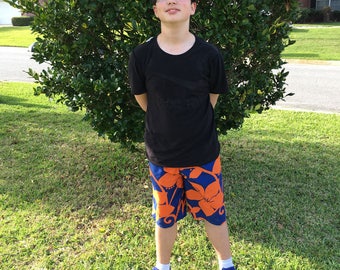 Pode incluir: Uma pessoa jovem vestindo uma camiseta preta e sunga azul e laranja com padrão floral. A pessoa está em pé na frente de um arbusto verde.