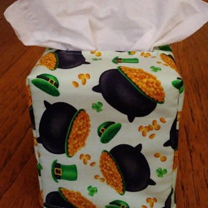 May include: A fabric tissue box cover featuring a St. Patrick's Day theme. The cover is light green with a repeating pattern of black pots of gold, green leprechaun hats, gold coins, and green shamrocks. White tissues are visible at the top.