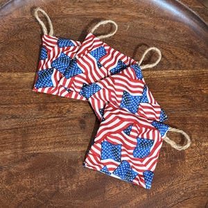 May include: Several small fabric pouches with a patriotic American flag pattern of red and white stripes and blue stars. Each pouch has a loop of natural twine attached for hanging. The pouches are arranged on a dark wooden tray.