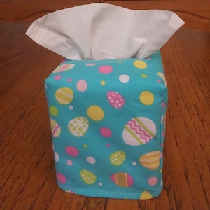 May include: A turquoise blue tissue box cover with a colorful Easter egg pattern. The cover has pink, yellow, green, and white eggs on a blue background.