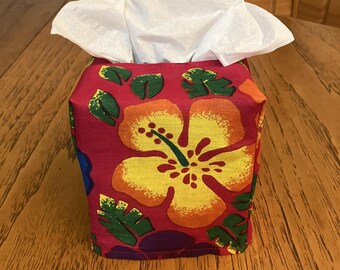 May include: A square fabric tissue box cover featuring a vibrant tropical pattern with large yellow and orange hibiscus flowers and green palm leaves on a deep magenta background, with a white tissue protruding from the top opening.