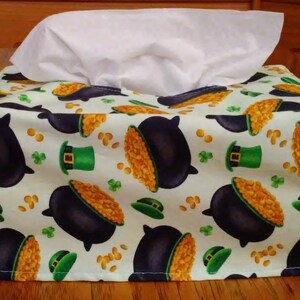 May include: A white tissue box cover with a green, gold, and black pattern of pots of gold and leprechaun hats.