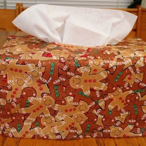 May include: A gingerbread man-themed tissue box cover. The cover is made of fabric and features a repeating pattern of gingerbread men in various poses. The tissue box is not included.