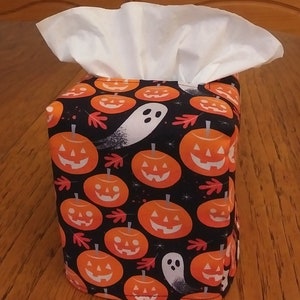 May include: A black tissue box cover with a Halloween theme. The cover features a repeating pattern of orange pumpkins with white faces and black stems on a black background. There are also white ghosts with black eyes on the cover.
