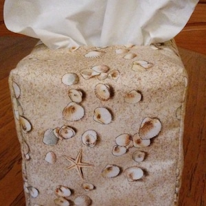 May include: A beige tissue box cover with a seashell and starfish pattern. The cover is made of fabric and has a white tissue box inside.