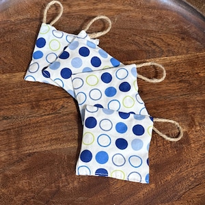 May include: Four square fabric coasters with a white background and blue and green polka dots. Each coaster has a loop of natural twine for hanging. The coasters are arranged on a dark wooden tray.