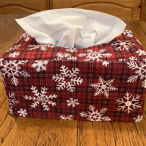 May include: A rectangular fabric tissue box cover featuring a red and black plaid pattern with white snowflake prints. The cover is designed to fit over a standard tissue box, with a central opening at the top where a white tissue is visible.