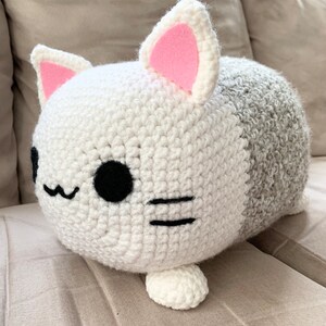 Può includere: Un cuscino a forma di gatto in crochet bianco e grigio con orecchie in feltro rosa e occhi e baffi ricamati neri.