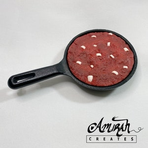 Peut inclure: Un cookie rouge et blanc dans une poêle en fonte noire. Le cookie est garni de pépites de chocolat blanc.