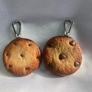 Peut inclure: Deux porte-clés en forme de cookie aux pépites de chocolat. Les cookies sont bruns avec des pépites de chocolat et ont une texture réaliste. Les porte-clés sont attachés à des boucles en métal argenté.