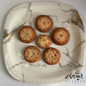May include: Six chocolate chip biscuits on a white marble-effect plate. The biscuits are round, with a light brown colour and small, dark chocolate chips. The plate is square with rounded corners and gold veining.