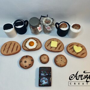 以下が含まれることがあります： チョコレートバー、クッキー、バターを塗ったトースト、卵を乗せたトースト、ハート型のバターを塗ったトーストなど、ミニチュアの食品のコレクションです。クッキーが入ったマグカップが2つと、クッキーが入った瓶が2つもあります。