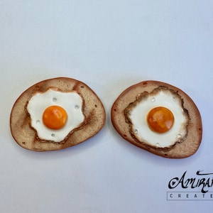 May include: Two brown clay ornaments shaped like fried eggs with orange yolks. The ornaments are on a white background.