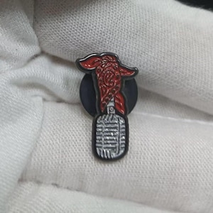May include: A red and black enamel pin featuring a stylized microphone with a red design on top. The pin has a black circular base and is likely a collectible or accessory.