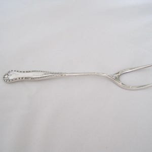 May include: A silver serving fork with a decorative handle and two tines. The handle has an ornate design, and the fork is made of a shiny, reflective metal. The fork is designed for serving food.