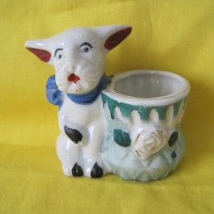 May include: A vintage ceramic planter shaped like a white dog with red ears and a blue scarf. The planter features a green and white floral design with a rose. The dog has black spots on its paws. A charming decorative item.