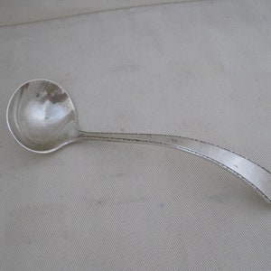 May include: A silver ladle with a rounded bowl and a long, curved handle. The handle has a decorative edge. The ladle is designed for serving soups, sauces, or other liquids. The ladle is made of a shiny, reflective metal.