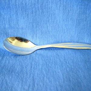 Puede incluir: Una cuchara plateada con un mango curvo y un cuenco redondeado. El mango presenta un diseño decorativo de dos líneas paralelas. La cuchara está sobre un fondo azul.