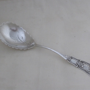Peut inclure: Une cuillère de service en argent avec un bol festonné et un manche détaillé. Le bol présente un motif floral gravé. La poignée présente un motif floral et des volutes. La cuillère est probablement en argent sterling.