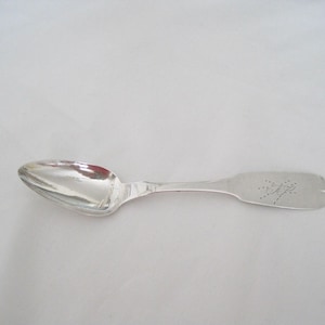 May include: A silver spoon with a rounded bowl and a long, flat handle. The handle has an engraved design near the end. The spoon is set against a white background.